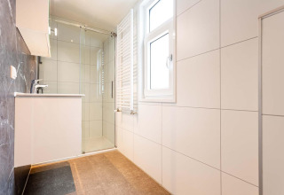 Cuarto de baño moderno con ducha de cristal, ventana grande y azulejos blancos en Deluxe Sauna de EuroParcs Noordwijkse Duinen.