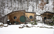 Casa Hobbit cubierta de nieve en el bosque de Limburg, Bélgica, con puerta redonda verde y detalles rústicos.
