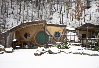 Casa Hobbit cubierta de nieve en el bosque de Limburg, Bélgica, con puerta redonda verde y detalles rústicos.
