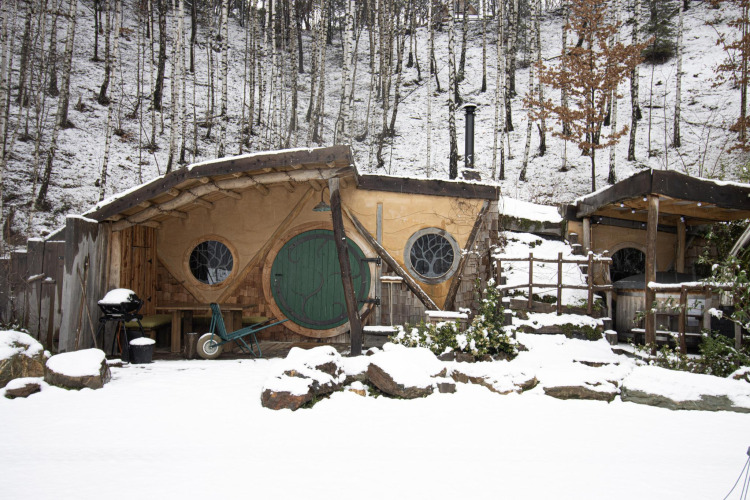 Casa Hobbit cubierta de nieve en el bosque de Limburg, Bélgica, con puerta redonda verde y detalles rústicos.