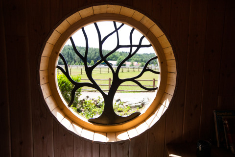 Fenêtre ronde avec motif d'arbre à Hobbit House 1, Cosy Cabins, donnant sur la forêt en Belgique.