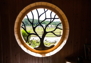 Round window with tree design in Hobbit House 1, Cosy Cabins, overlooking forest landscape in Belgium.