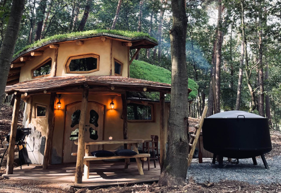 Sprookjesachtige Hobbit House lodge in het bos met grasdak, houten terras en natuurlijke sfeer.