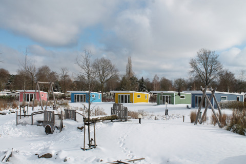 Bunte Tiny Houses im Schnee bei Valley Chalet, Holiday park Ackersate, Niederlande, mit Spielplatz vorne.