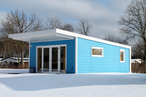Valley Chalet, una tiny house azul en la nieve en Holiday park Ackersate en Países Bajos, rodeada de árboles.