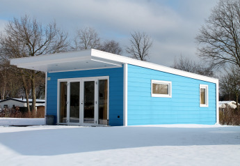 Valley Chalet, een blauw tiny house in de sneeuw bij Holiday park Ackersate, Nederland, omgeven door bomen.
