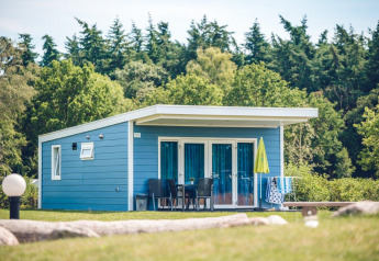 Blauwe tiny house Valley Chalet op vakantiepark Ackersate in Nederland, omgeven door bomen en groen gras.