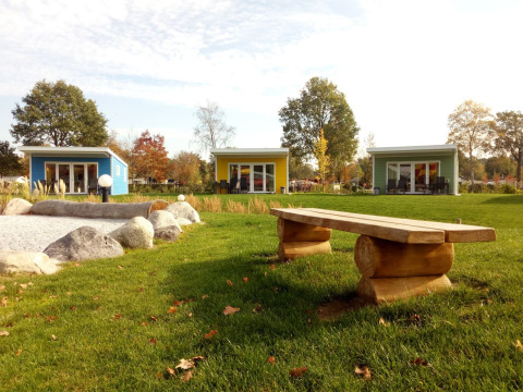 Bunte Valley Chalet Tiny Houses im Holiday Park Ackersate, Niederlande, mit Wiese, Steinen und Holzbank.