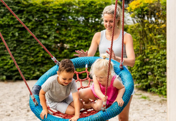 Eine Frau schubst zwei kleine Kinder auf einer Nestschaukel im Sand bei Wood Cottage, Ferienpark De Witte Berg.