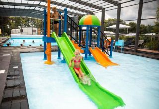 Children play on colorful water slides in an indoor pool area at a glamping accommodation in summer.