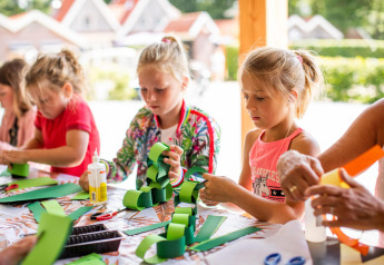 Bambini che fanno lavoretti di carta seduti a un tavolo durante una vacanza in glamping al sole.