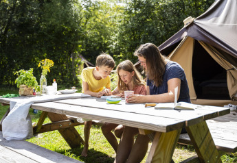 Familie sitzt an einem Picknicktisch vor einem Safarizelt bei Glamptent GT, BreeBronne, Niederlande, und genießt die Natur.