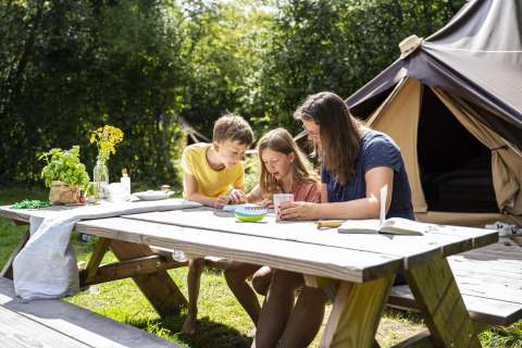 Familie sitzt an einem Picknicktisch vor einem Safarizelt bei Glamptent GT, BreeBronne, Niederlande, und genießt die Natur.