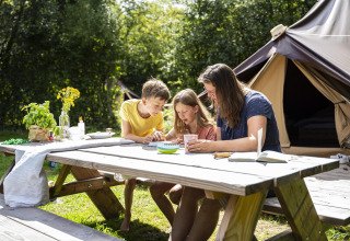 Familia disfruta en una mesa de picnic frente a una tienda en Glamptent GT, BreeBronne, Países Bajos, rodeados de naturaleza.