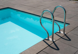 Udendørs swimmingpool med rent vand og stålrækværk ved et glampingophold, omgivet af flisebelægning.