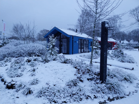 Cabaña de caza con sauna cubierta de nieve en el parque Holiday Ackersate, Países Bajos, rodeada de árboles.