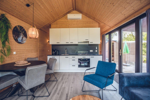Intérieur de lodge moderne avec cuisine ouverte, coin repas, bois et grandes fenêtres donnant sur la terrasse.