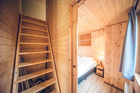 Interior de una cabaña revestida de madera con una sencilla escalera y dormitorio con cama doble y mesita de noche.