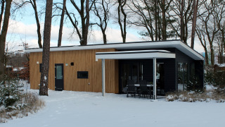 Moderne Forest Villa lodge in de sneeuw bij Vakantiepark Ackersate, Nederland, omgeven door hoge bomen.