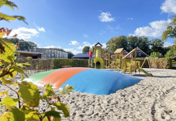 Legeplads med hoppepude, rutsjebaner og legeredskaber på en glampingplads under en klar blå himmel.