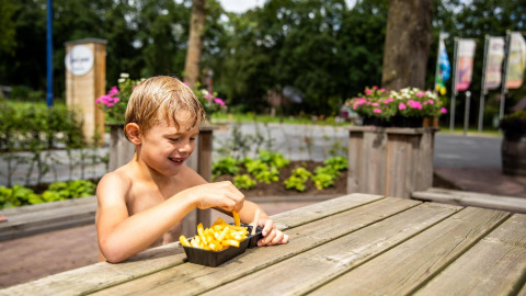 Niño comiendo papas fritas en una mesa de madera junto a la tienda Tentvilla en el parque De Witte Berg.