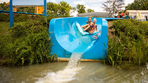 Deux enfants glissent sur un toboggan bleu dans un étang, sur un site de glamping avec tentes en arrière-plan.
