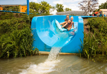 Two children ride a blue waterslide into a pond at a countryside glamping site with tents in view.