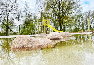 Steine im Wasser vor einem Spielplatz mit Rutsche bei Forest Lodge, De Witte Berg, Niederlande, Frühling.