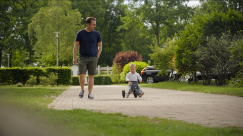 A man walks beside a young child riding a tricycle at Forest Lodge, De Witte Berg holiday park, Netherlands.