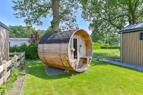 Sauna de madera en forma de barril junto a un lodge de lujo en Drentse Weelde, Países Bajos, rodeado de vegetación.