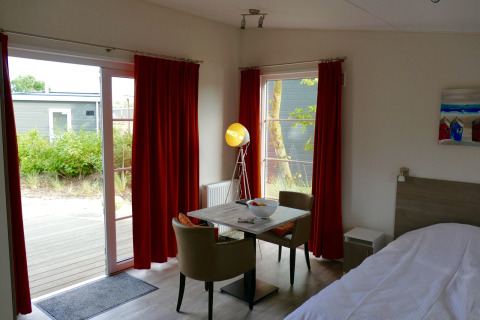 Interior de la tiny house Comfort Lodge en Kompas Camping Nieuwpoort, Bélgica, con mesa y cortinas rojas.
