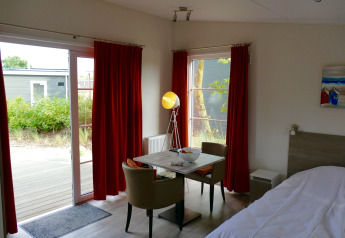 Interior de la tiny house Comfort Lodge en Kompas Camping Nieuwpoort, Bélgica, con mesa y cortinas rojas.