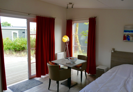 Interno della tiny house Comfort Lodge a Kompas Camping Nieuwpoort, Belgio, con tavolo e tende rosse.