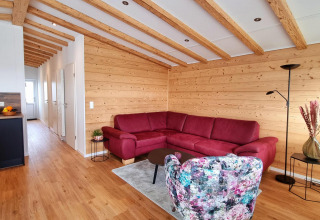 Sala en Chalet Enzian con paredes de madera, vigas, sofá rojo y silla floral en Camping & Ferienpark Brilon, Alemania.