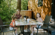A family sits at an outdoor table at Happy bee lodge, Camping Si-Es-An in the Netherlands, surrounded by trees.