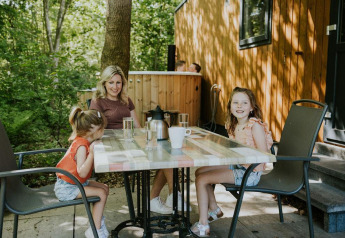 En familie sidder ved et bord udenfor Happy bee lodge på Camping Si-Es-An, omgivet af grønne træer.