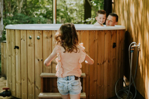 Une enfant marche vers un jacuzzi extérieur où un homme et un garçon sourient, entourés de bois et de verdure.