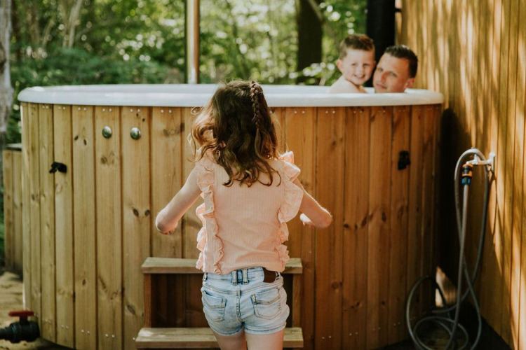 Una niña se acerca a un jacuzzi exterior donde un hombre y un niño sonríen, rodeados de paneles de madera y árboles.