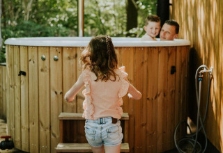 Una niña se acerca a un jacuzzi exterior donde un hombre y un niño sonríen, rodeados de paneles de madera y árboles.