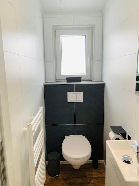 Baño moderno blanco con inodoro suspendido, ventana pequeña y lavabo en Chalet Bergrose, Alemania.