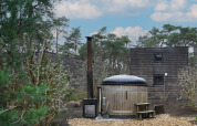 Wooden tiny house with hot tub and chimney among trees in Beekbergen, Netherlands under blue sky.