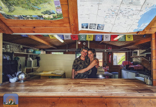 Een glimlachend koppel omhelst elkaar achter een houten toog in Camping Des Randonneurs, Occitanië, Frankrijk.