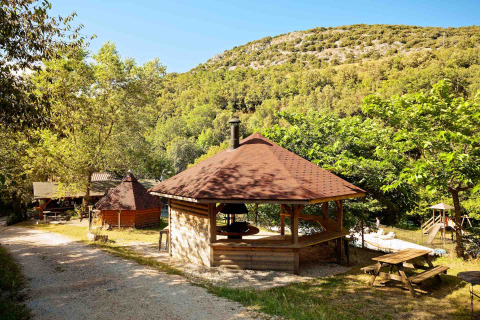 Camping Des Randonneurs in Occitania, Francia, con gazebo barbecue, tavoli da picnic e colline verdi sullo sfondo.