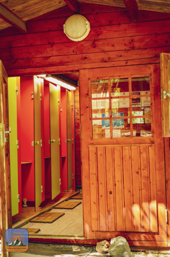 Entrée d’une cabane en bois avec portes ouvertes, révélant des cabines de toilettes colorées en glamping.