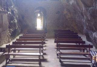 Stone benches inside a cave chapel near Fenouillet, Occitanie, France, with sunlight at the entrance.