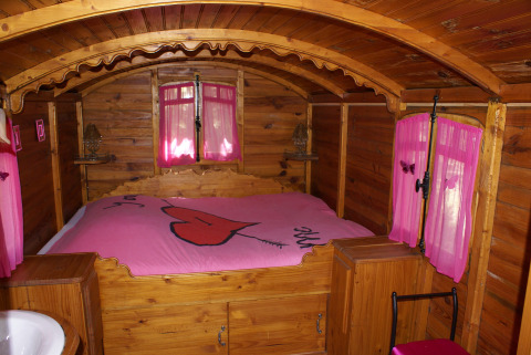 Interior de una acogedora cabaña de madera con cortinas rosas y una cama con colcha decorada con un gran corazón.