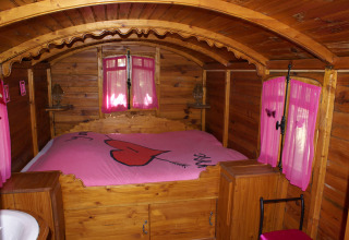 Interior de una acogedora cabaña de madera con cortinas rosas y una cama con colcha decorada con un gran corazón.