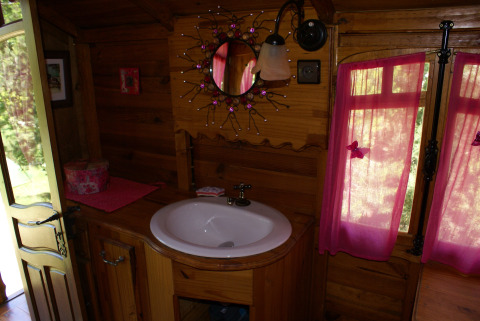 Bagno in legno accogliente con lavandino rotondo, specchio decorato e tende rosa in una roulotte gipsy in Francia.