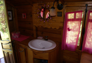 Acogedor baño de madera con lavabo redondo, espejo decorativo y cortinas rosas en una carreta gitana en Francia.
