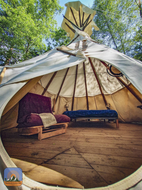 Interior de una tienda tipi Wigwam con suelo de madera, cama y sofá en Camping Des Randonneurs, Francia.